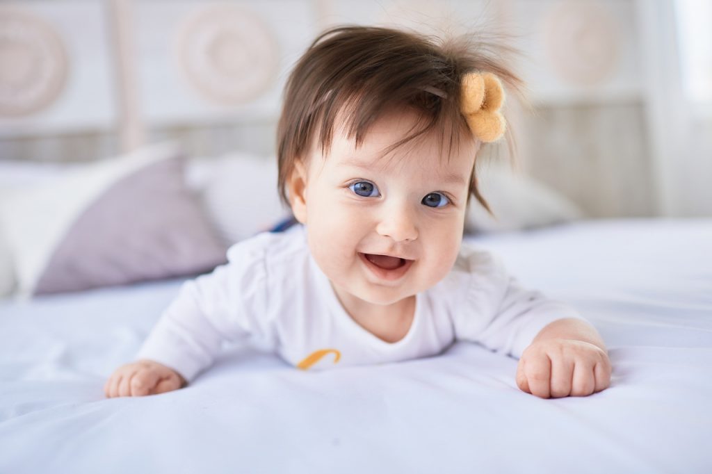 bebê com roupa branca deitada, com a cabeça erguida, em uma cama, com laço laranja no cabelo