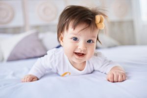 bebê com roupa branca deitada, com a cabeça erguida, em uma cama, com laço laranja no cabelo